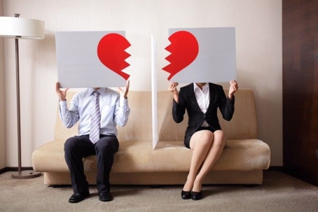 Two people holding signs with a half a broken heart over their faces sitting on a couch with a barrier between them.