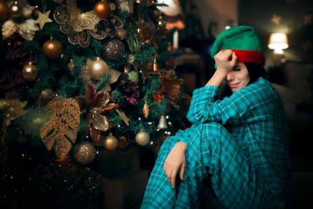 A person in pajamas and an elf cap sitting next to a Christmas tree crying.