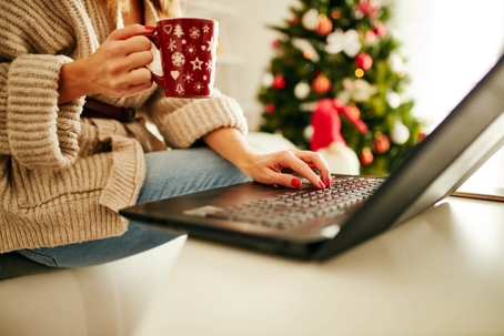 Woman holding cup of coffee while working on laptop at home for Christmas.