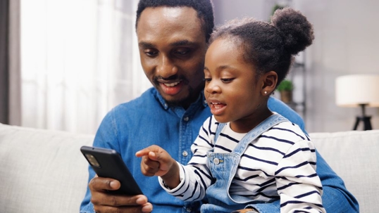 A father and daughter sitting on the couch playing with a smartphone.